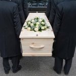 Four people carrying a coffin with white flowers.