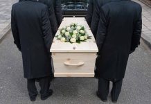 Four people carrying a coffin with white flowers.