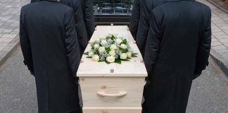 Four people carrying a coffin with white flowers.