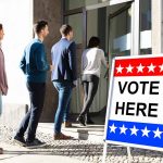 People entering building with "Vote Here" sign nearby.