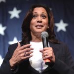 Woman speaking with microphone, American flag background.