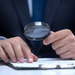 Man holding magnifying glass, examining document on clipboard.