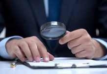 Man holding magnifying glass, examining document on clipboard.